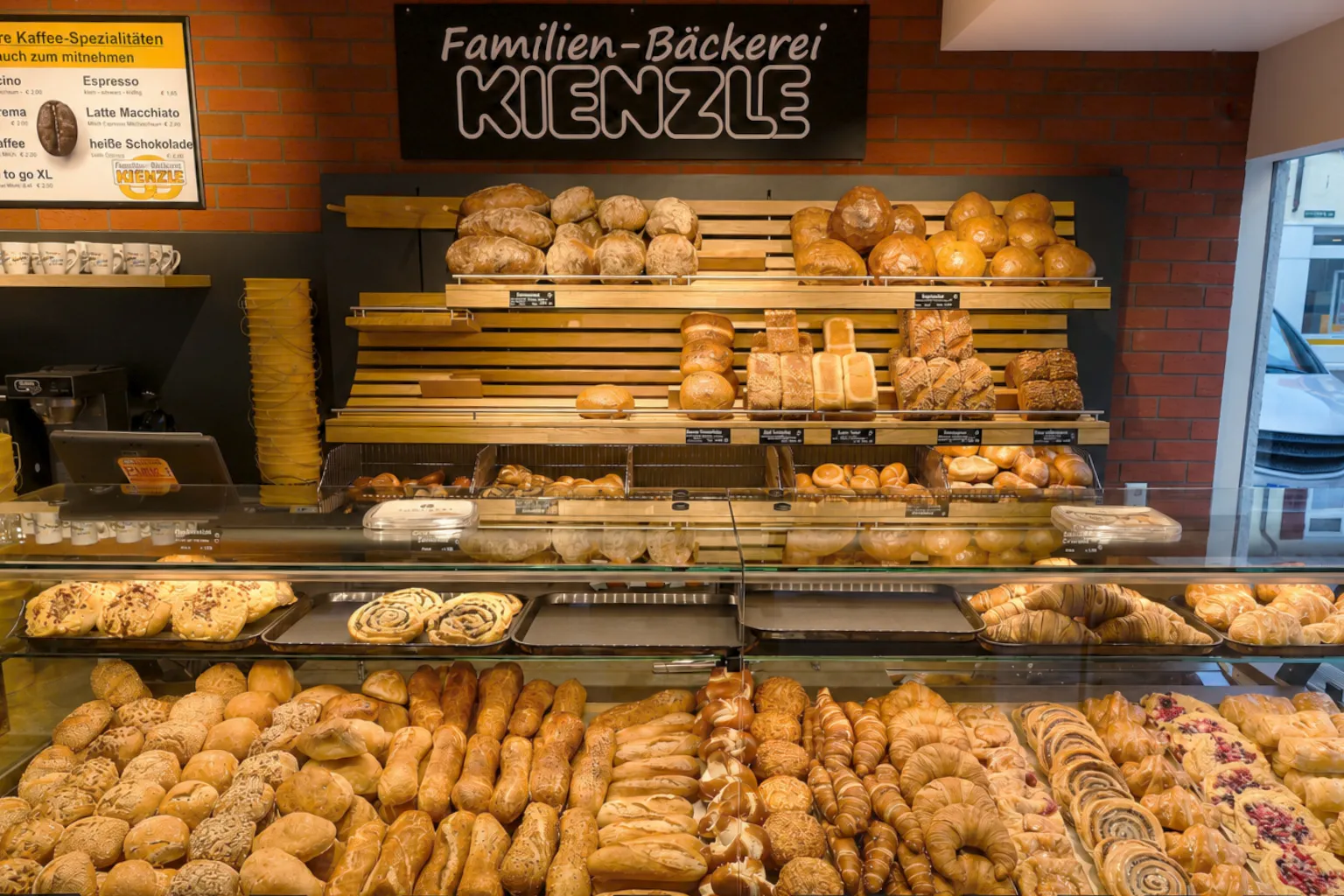 Die Verkaufstheke im Stammhaus Gaußstraße: eine gefüllte Auslage mit Brötchen, Hörnchen und Teilchen, darüber die Regale mit Broten und Laugengebäck, links die Kaffeemaschine mit der Karte der Kaffee-Spezialitäten, darüber das Schild „Familien-Bäckerei Kienzle“